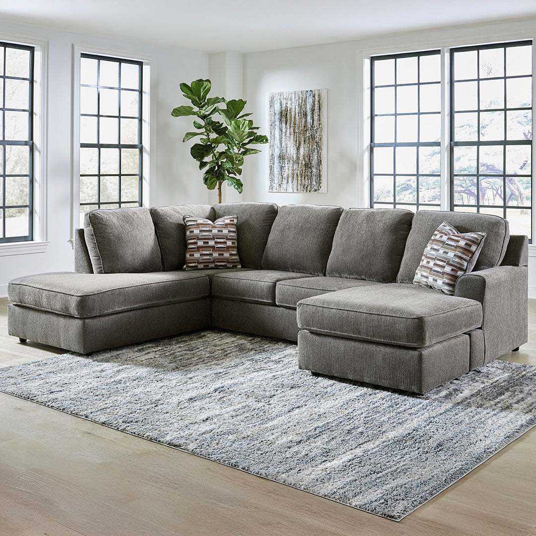 Gray sectional sofa with patterned pillows on a rug in a room with large windows.