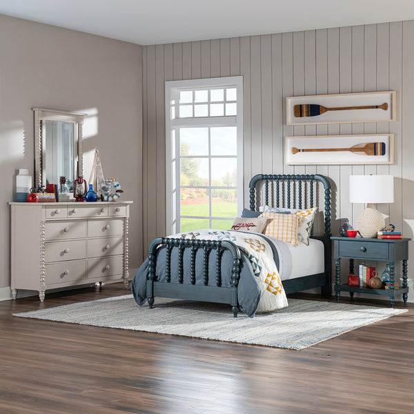 A styled bedroom scene showing a dark blue wooden bed frame with a decorative headboard and footboard, alongside a matching blue nightstand and a cream-colored dresser with a mirror.