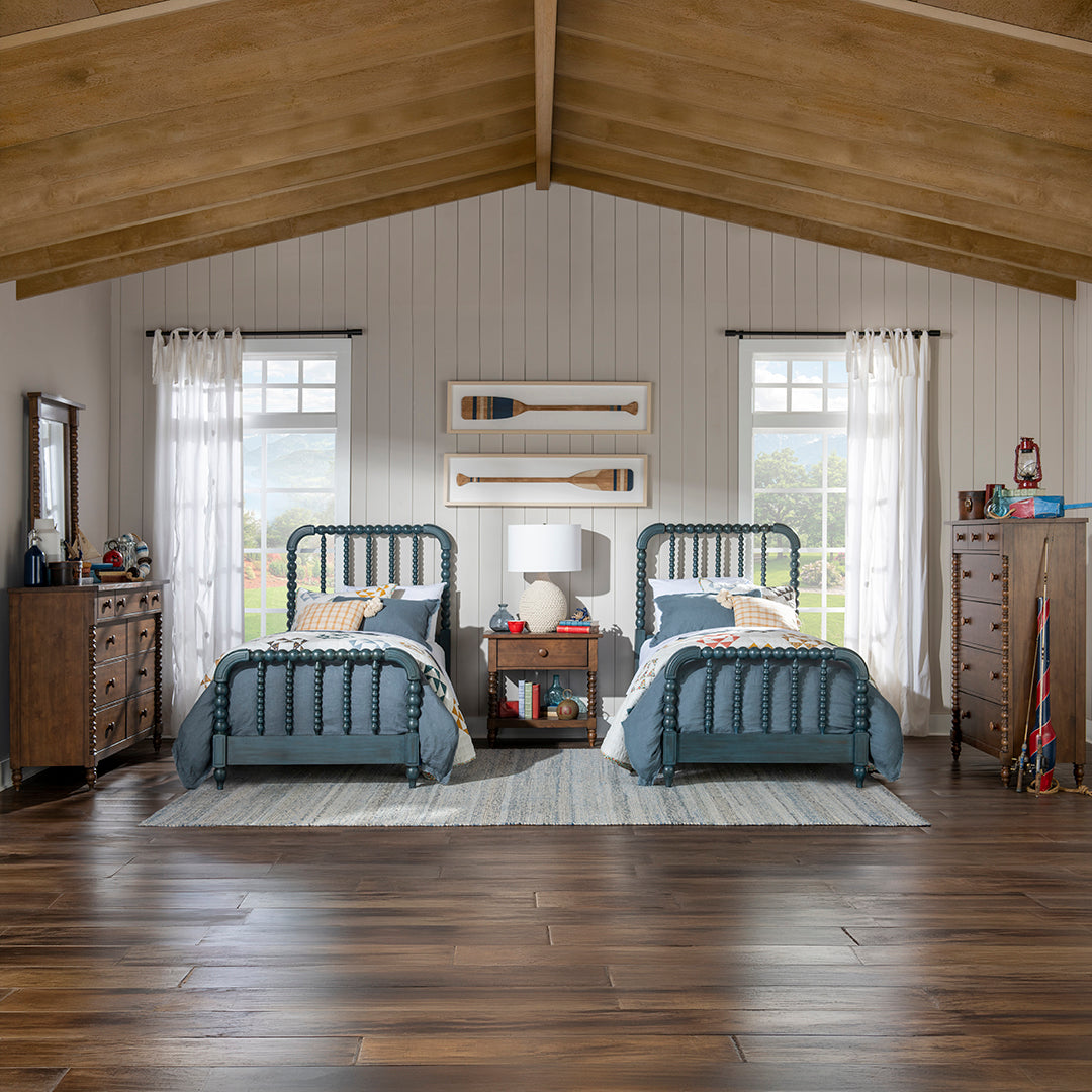 A styled bedroom interior showing two matching blue metal twin beds with blue bedding, flanking a central nightstand and window, in a rustic room with wood floors, paneled walls, and a high wood-beamed ceiling.