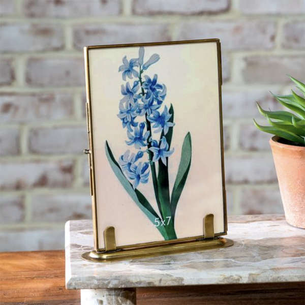 A vertical 5x7 inch picture frame with a gold-toned metal border and stand, featuring a blue hyacinth floral print on a cream background, displayed on a marble-topped table with a brick wall and potted plant in the background.