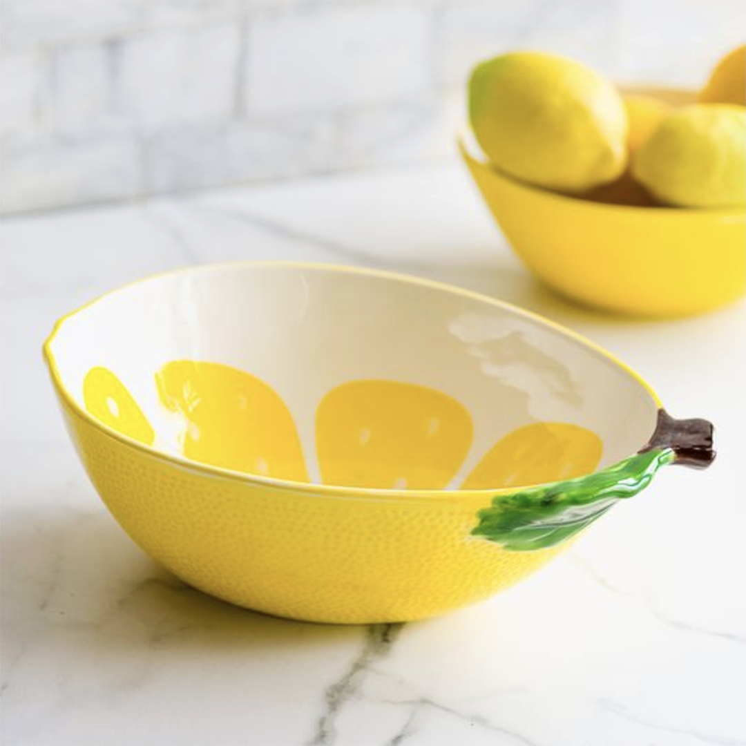 A yellow lemon-shaped bowl with a textured exterior, a white interior decorated with yellow lemon slices, and a green leaf and brown stem detail on the side, set on a marble countertop with a blurred yellow bowl of lemons in the background.