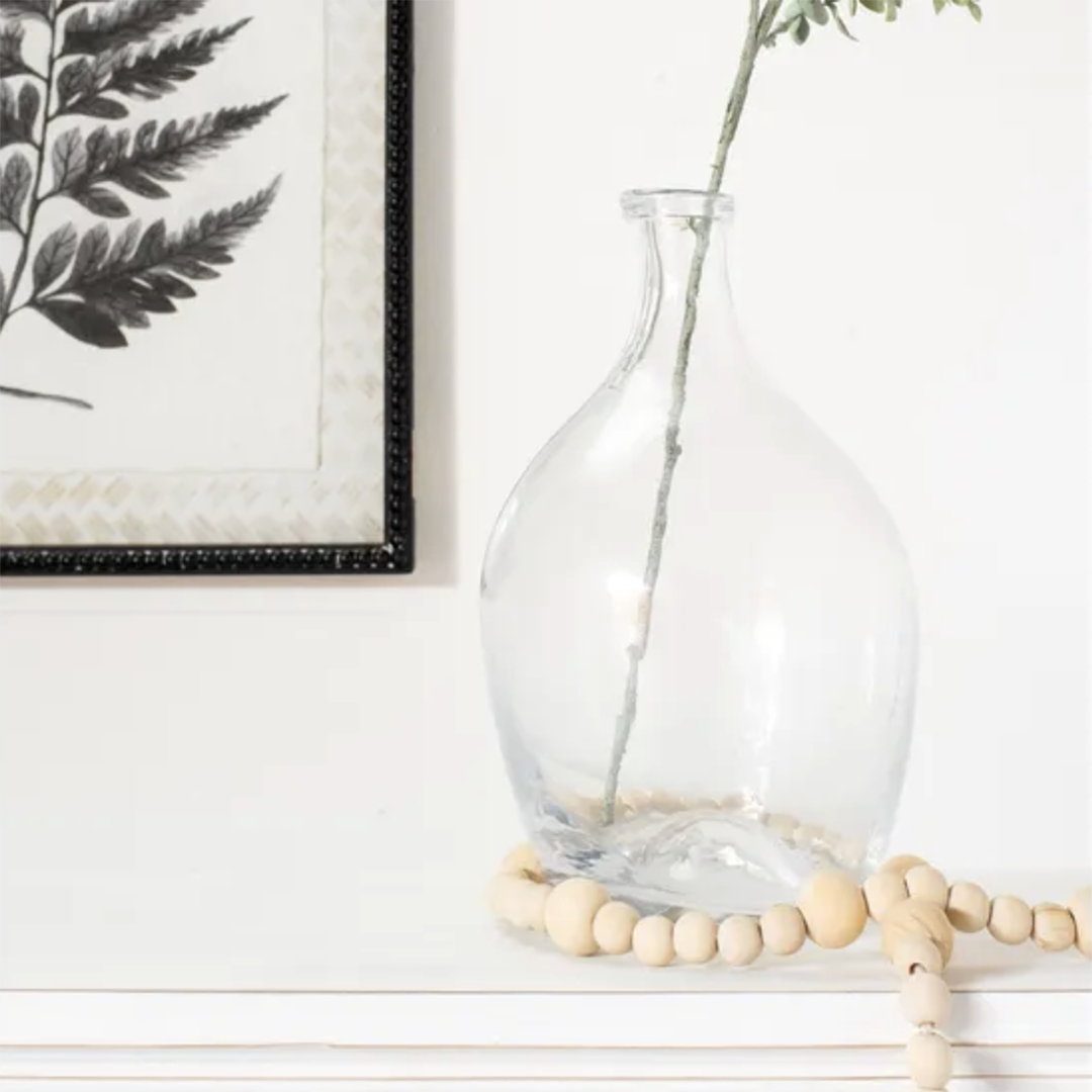 Clear glass vase with a rounded bulbous body and narrow neck, holding a long green stem, styled on a white surface with a wooden bead garland and a framed botanical print in the background.