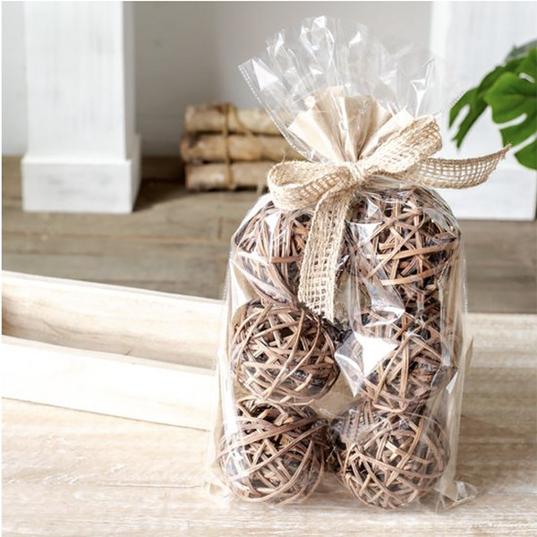 A clear cellophane gift bag containing several natural woven rattan balls, tied with a beige burlap ribbon, set on a light wood surface in a softly blurred indoor setting.