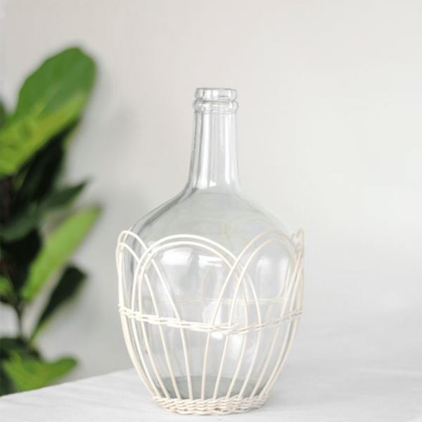 A clear glass bottle with a wide rounded body and narrow neck, wrapped in a cream-colored woven wire cage, set on a white surface with a blurred green plant in the background.