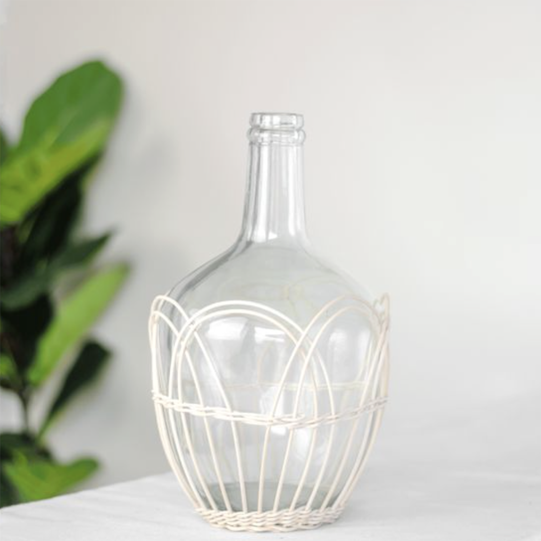 A clear glass bottle with a wide rounded body and narrow neck, wrapped in a cream-colored woven wire cage, set on a white surface with a blurred green plant in the background.
