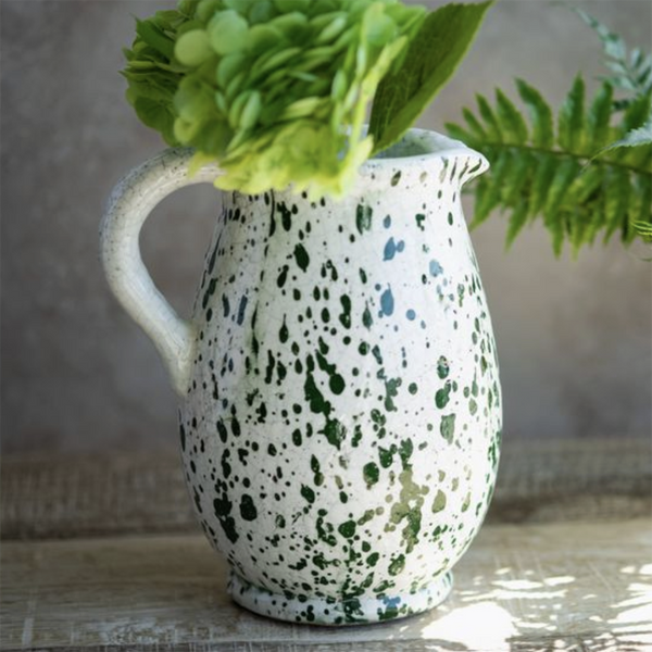 A white ceramic pitcher with green speckled glaze, filled with green hydrangea blooms and fern leaves, set on a rustic wooden surface against a neutral background.