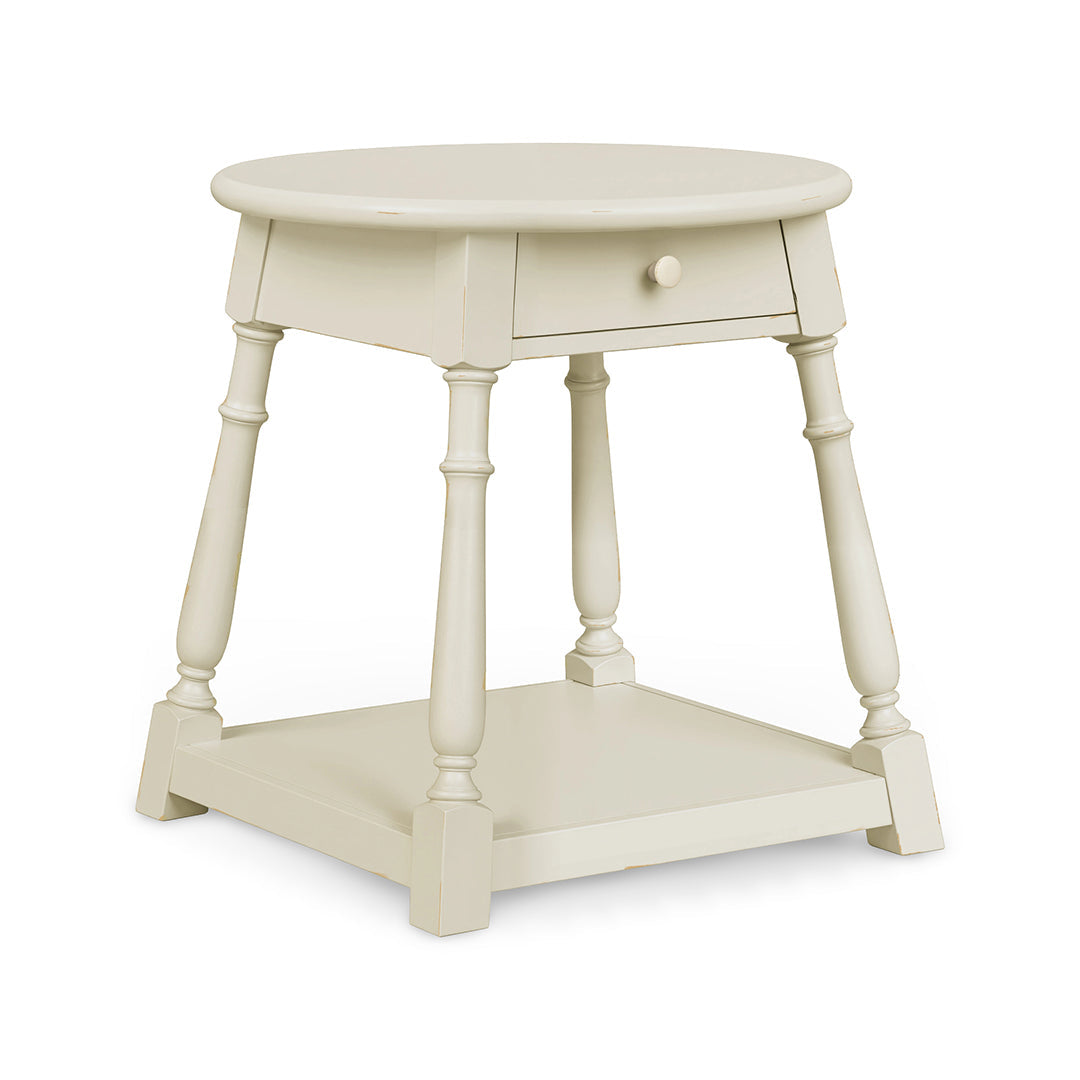A small round side table with a single drawer, turned legs, and a lower shelf, painted in a warm cream or off-white finish