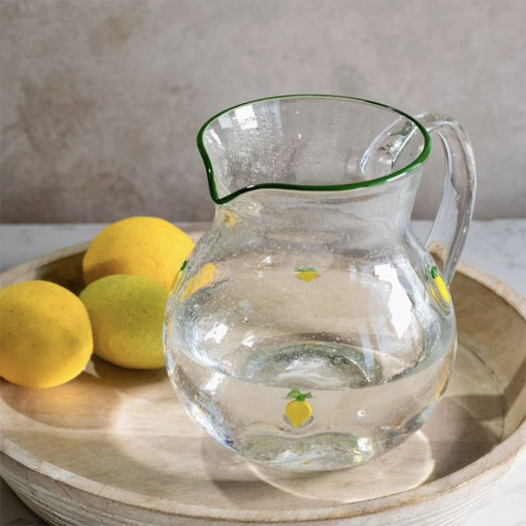 A clear glass pitcher with a green rim and small yellow lemon illustrations, filled with water and placed on a round wooden tray with lemons in the background.
