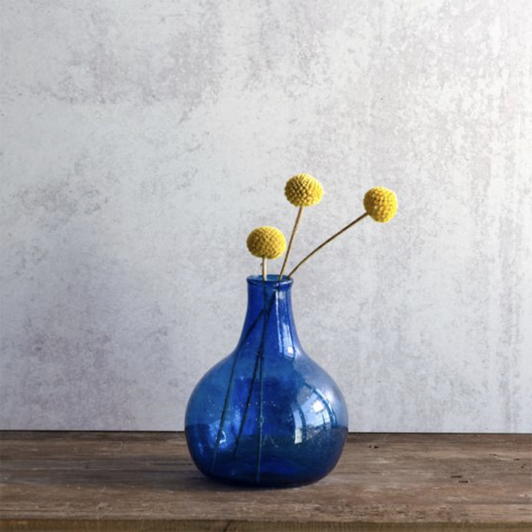 A cobalt blue glass vase with a rounded bulbous body and narrow neck, holding three yellow pom-pom flowers, set on a rustic wooden surface against a textured light gray wall.