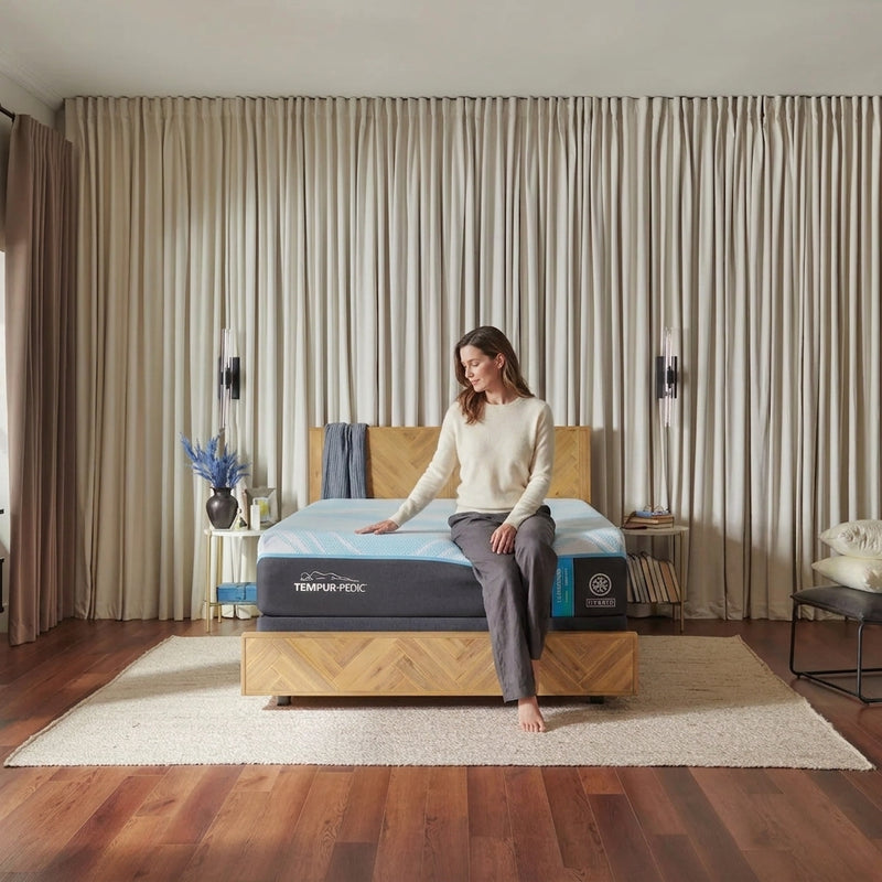 Woman in her early 40s sitting on the end of a bed, running her hand across the Tempur-Pedic LuxeBreeze Hybrid mattress to feel its softness