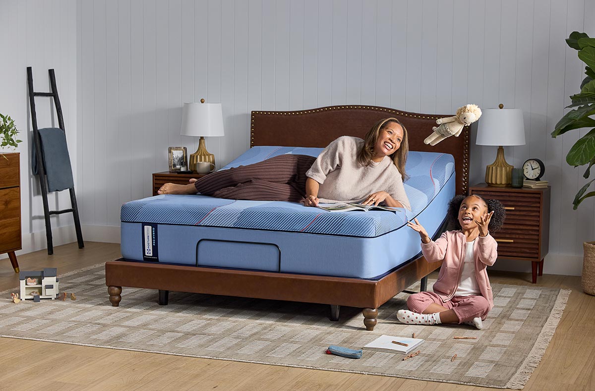 Woman reading on Sealy Posturepedic Pro Hybrid adjustable bed with child playing in bedroom