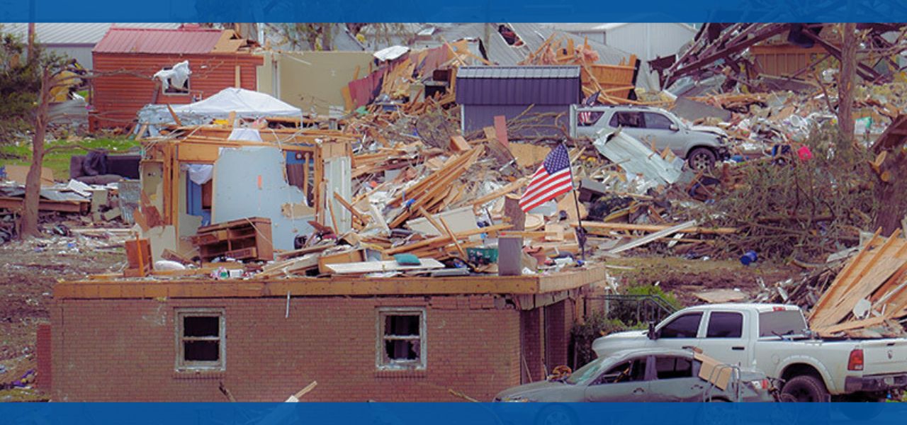 Damaged buildings and debris after a disaster, with a vehicle in the foreground.