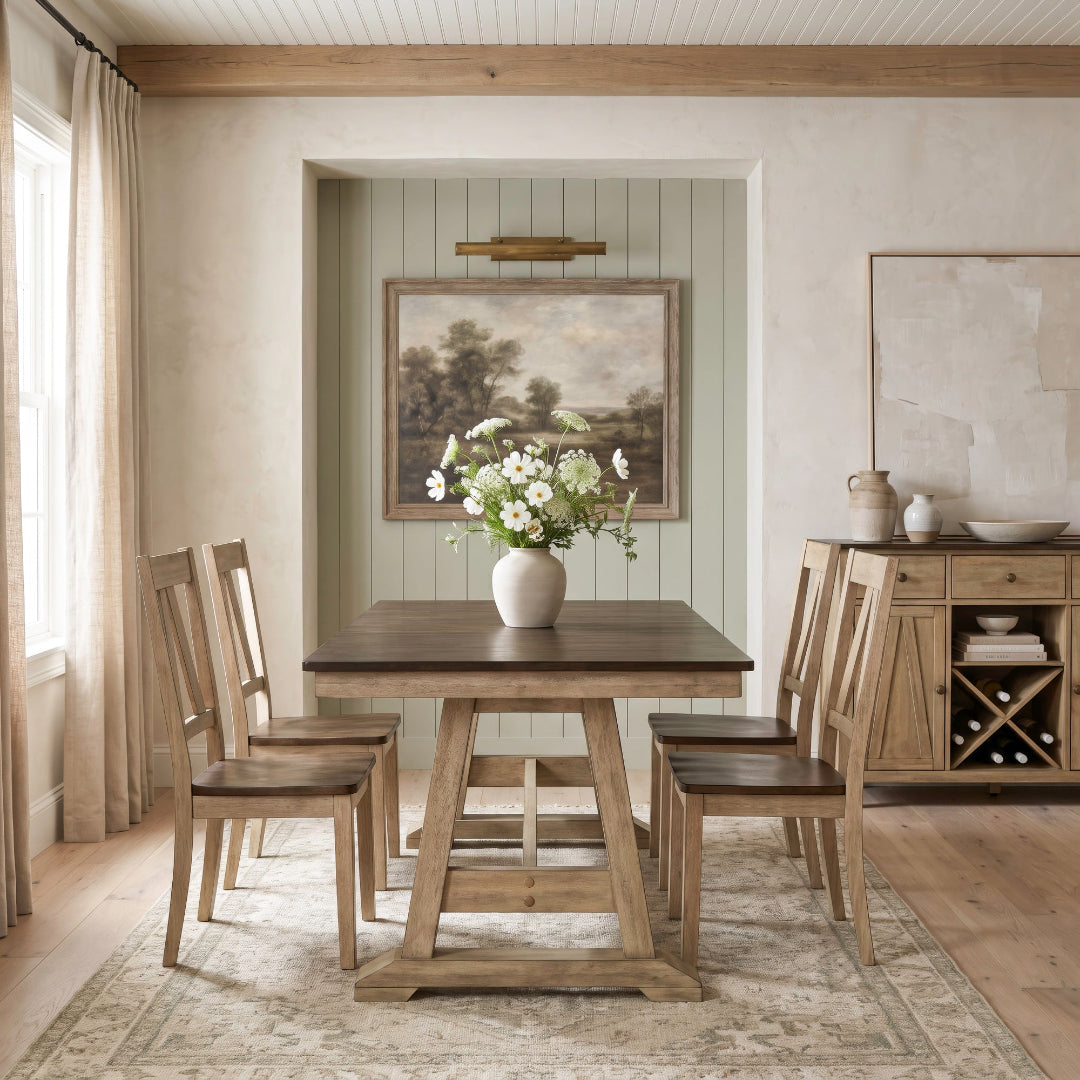 A rustic wooden dining table with four matching chairs in a warm, neutral-toned dining room. A white ceramic vase filled with white flowers sits on the table, which is placed on a patterned area rug.
