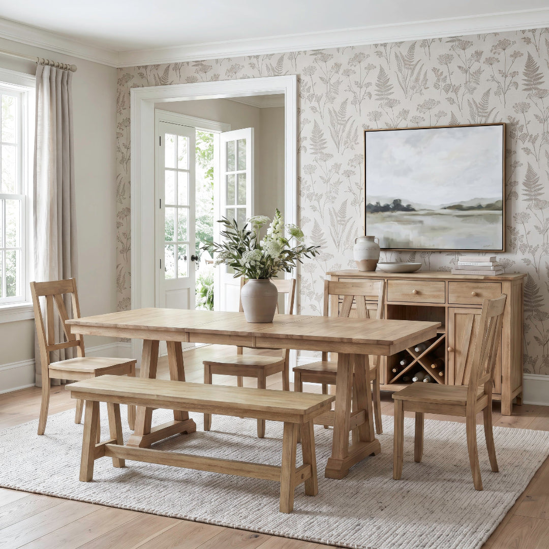 A rustic light oak dining room set featuring a rectangular wooden table, matching bench, and four chairs, styled in a bright dining room with botanical wallpaper and a sideboard.