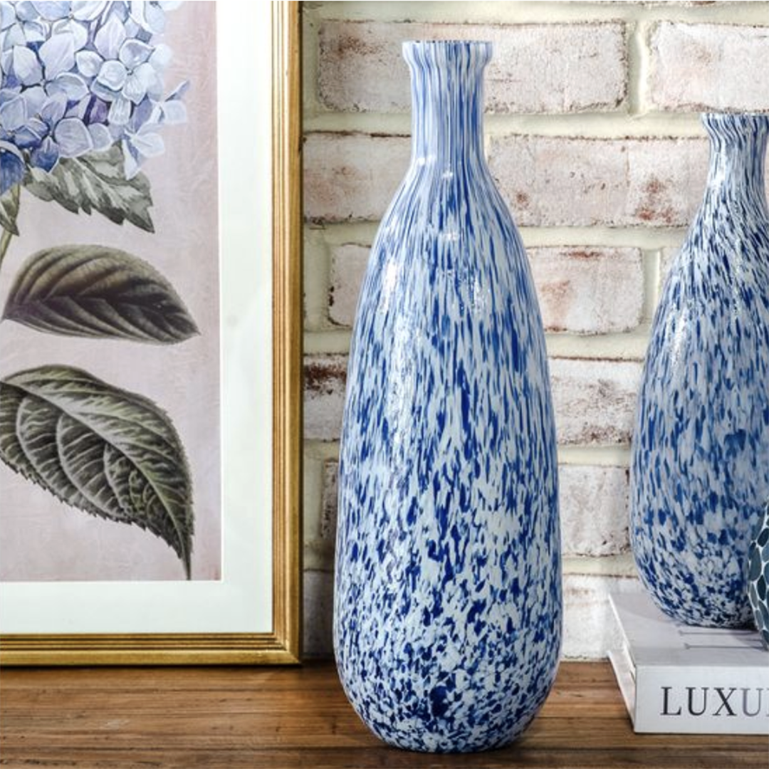 A tall blue and white speckled ceramic vase stands on a wooden surface against a white brick wall, with a framed botanical print partly visible on the left and another vase on a book to the right.