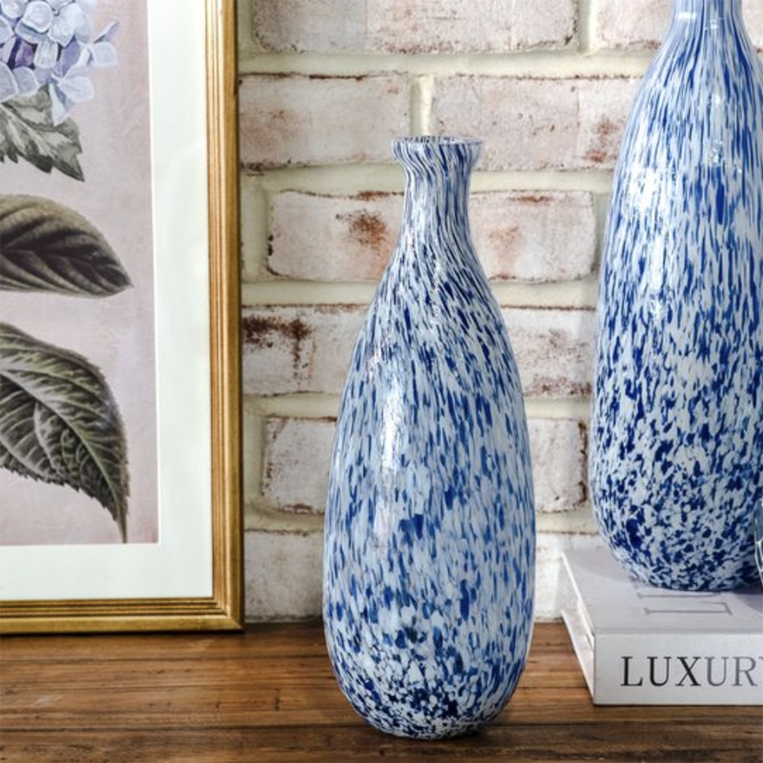 Two tall blue and white speckled ceramic vases displayed on a wooden shelf against a white brick wall, with a framed botanical print on the left and a white book on the right.