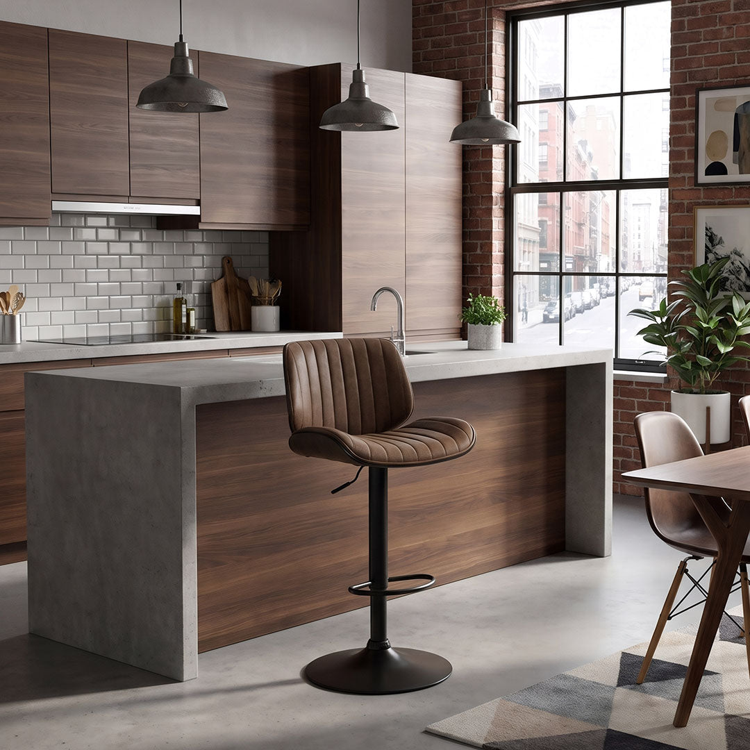 A modern kitchen island with a brown upholstered bar stool in a real kitchen environment, featuring wood cabinetry, concrete countertops, pendant lights, and a large window.