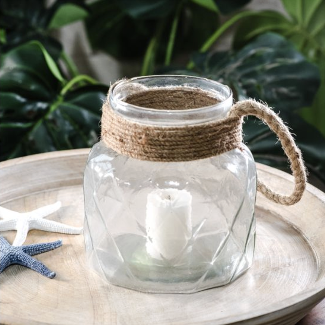 A clear faceted glass jar candle holder with a natural jute rope handle and collar, containing a white taper candle, displayed on a round wooden tray with starfish props and a blurred green plant background.