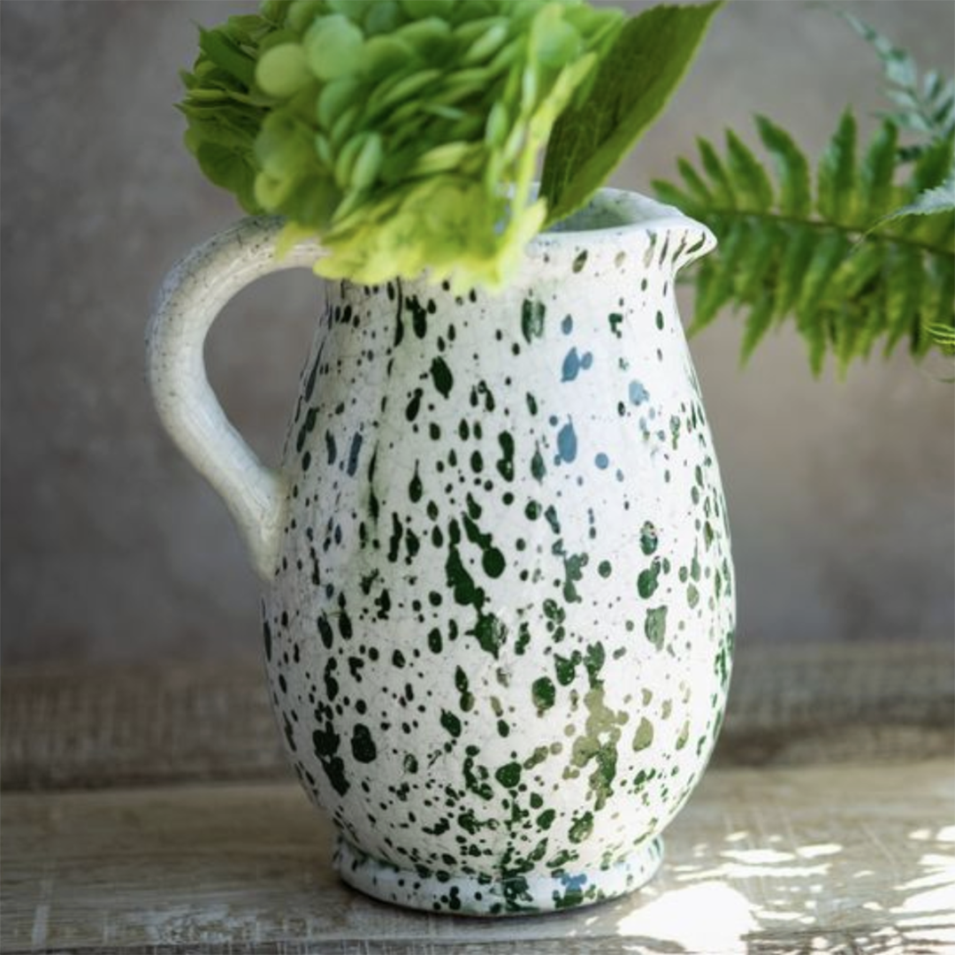 A white ceramic pitcher with green speckled glaze, filled with green hydrangea blooms and fern leaves, set on a rustic wooden surface against a neutral background.