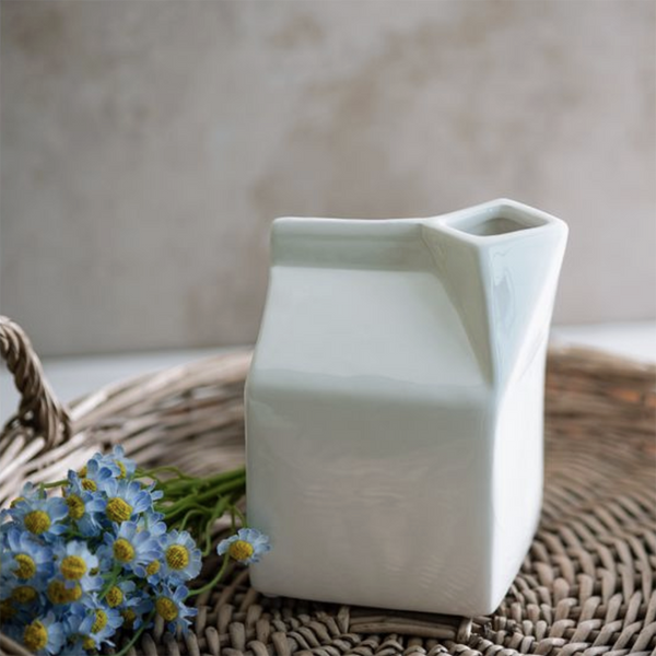 A matte white ceramic milk carton-shaped vase with a small opening at the top, styled on a woven wicker tray with blue daisies against a neutral beige background.