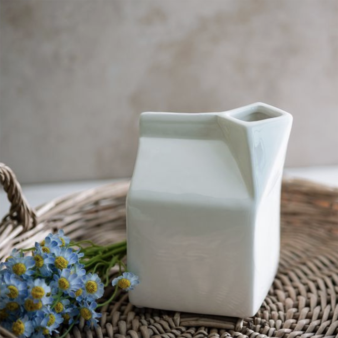A matte white ceramic milk carton-shaped vase with a small opening at the top, styled on a woven wicker tray with blue daisies against a neutral beige background.