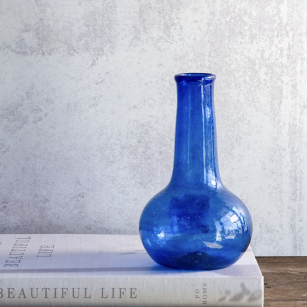 A cobalt blue glass vase with a bulbous base and narrow neck, displayed on top of a white book against a textured light gray wall.