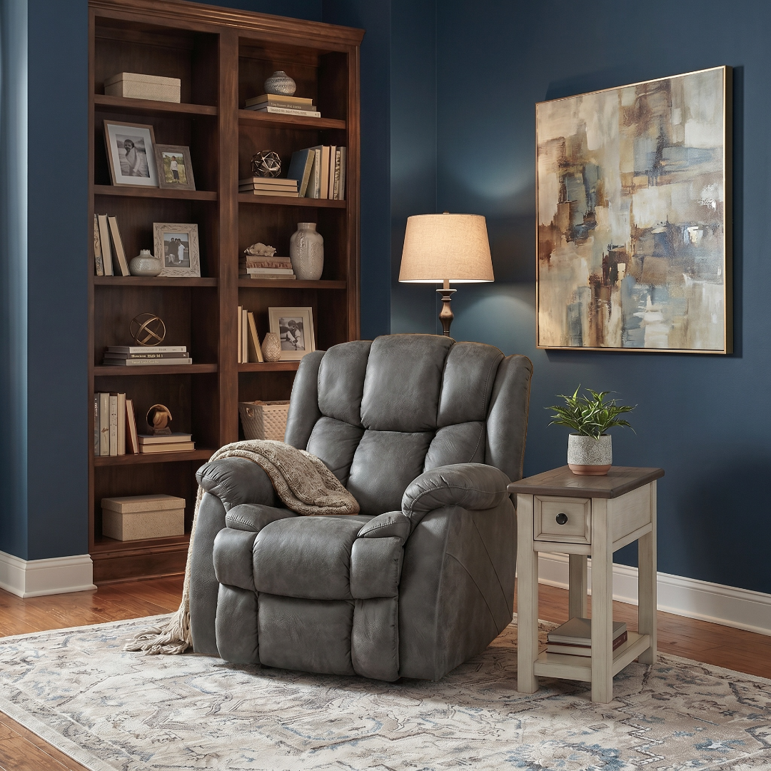 Gray recliner chair in a living room with bookshelf, lamp, and painting.