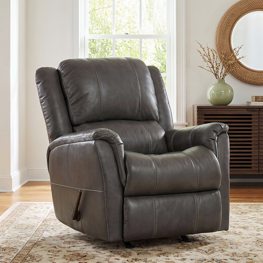 A dark gray faux leather recliner chair with tufted stitching, shown in a bright living room setting with a window, wooden cabinet, and patterned rug.