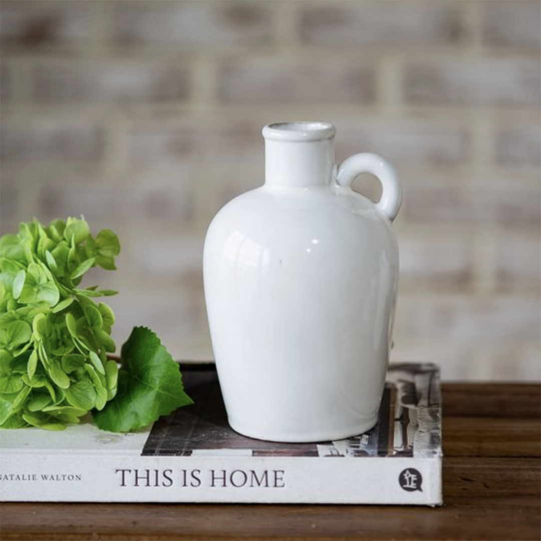 A glossy white ceramic jug-shaped vase with a small handle, displayed on top of a book with green hydrangea leaves in the foreground and a blurred brick wall background.