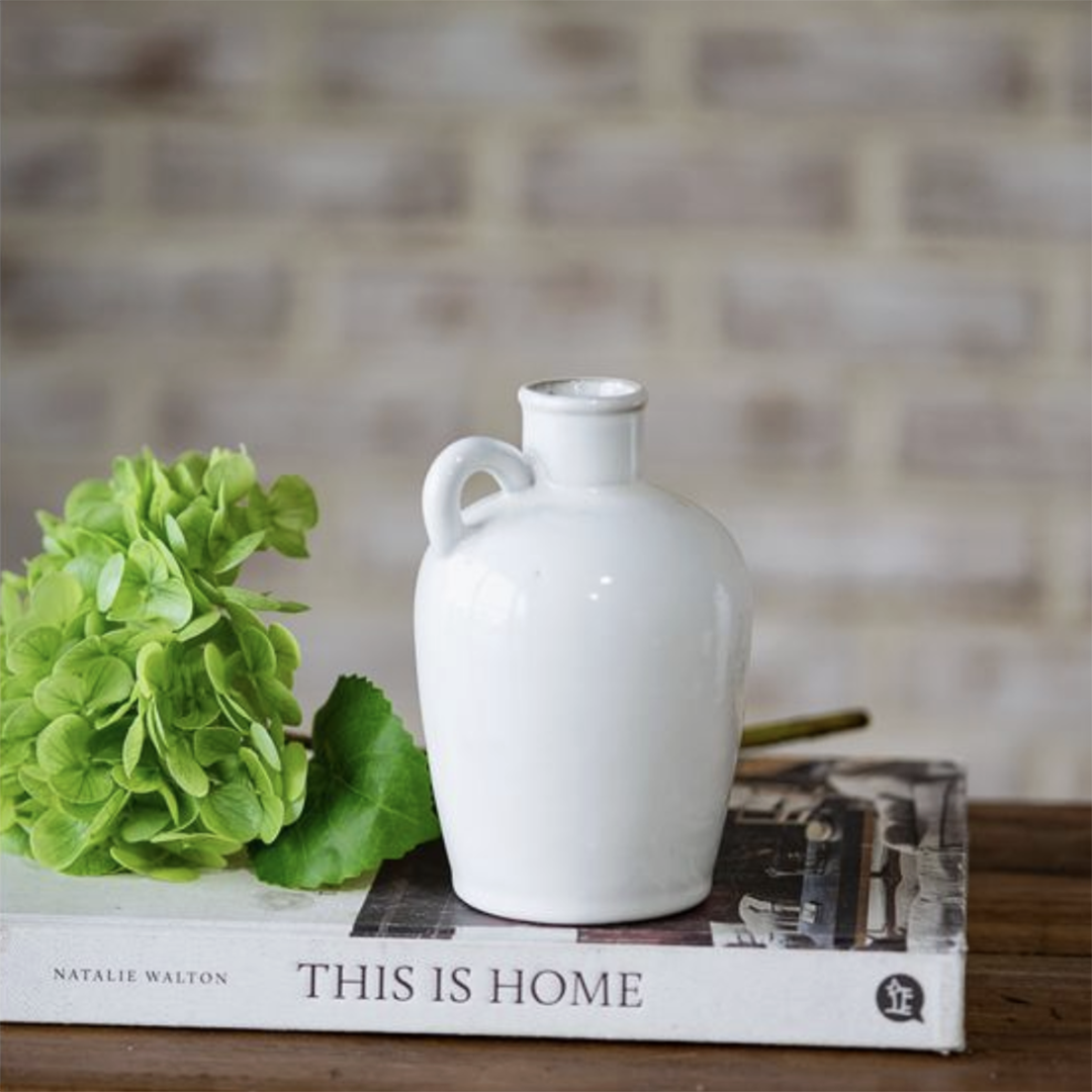 A glossy white ceramic jug-shaped vase with a handle, displayed on top of a book next to a green hydrangea stem against a blurred brick wall background.