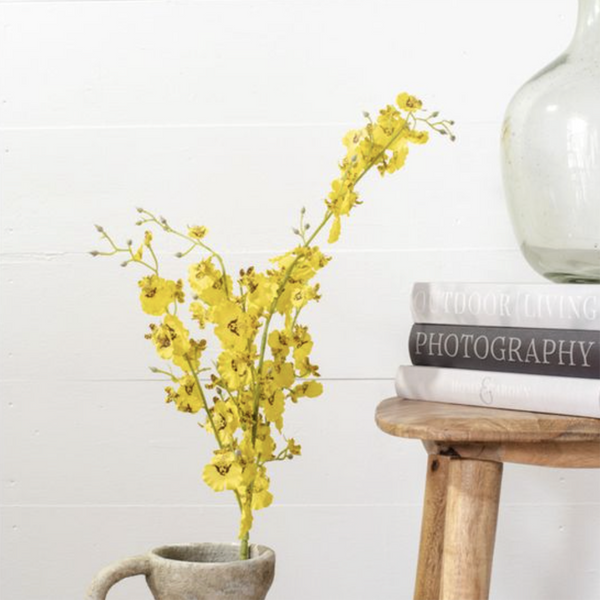 Yellow artificial orchid flowers arranged in a rustic gray ceramic mug, placed beside a small wooden table holding books and a clear glass bottle against a white shiplap wall.