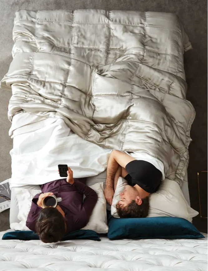 Overhead view of a couple in bed, one partner sleeping undisturbed while the other sits up with coffee