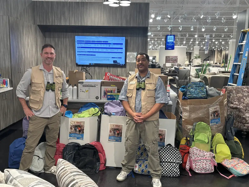 Greenwood store manager Chris Anastasio and sales associate Desmond Taylor in safari guide outfits from the Wild Weekend sale standing with donated backpacks and supply boxes